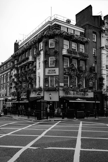 A black-and-white photograph of a multi-story building situated on a street corner, displaying several balconies adorned with hanging flower baskets and plants. The ground floor features a pub or restaurant with an awning and outdoor seating area enclosed by a railing. A few individuals are walking on the sidewalk nearby, and the street in front has parking restrictions marked by painted lines. The building's exterior showcases a mix of brick and concrete architectural elements, with a sign reading 'GRESHAM COLLECTIVE' attached to the side. The scene appears to be a typical urban setting in Paddington, London. This image can relate to house moves or relocations, illustrating the type of environment that might be involved in local removal services by Self Storage Paddington, supporting content on packing, loading, and transportation during a home relocation process.