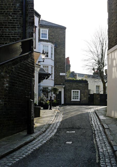 A street view capturing a residential area near Praed Street in Paddington, with a large multi-story brick building featuring decorative window frames and a steeply pitched roof with chimneys on the left side. The foreground shows a paved pavement with fallen leaves and a curved road with a curb, leading to a vanishing point where a small van is parked near the curb. Green trees with dense foliage overhang the roadway, partially shading the street, and a white wrought-iron fence lines the front of the building. The sky is overcast with grey clouds. This scene illustrates the typical exterior environment involved in house relocations and moving logistics in Paddington, as coordinated by Self Storage Paddington, including the preparation for a home moving process involving transportation, packing, and furniture transport along this quiet residential street.