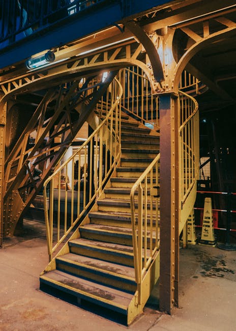 A red metal fire escape staircase attached to the exterior of a building, with multiple landings and angled steps leading up to the upper floors. The staircase features safety railings along the sides